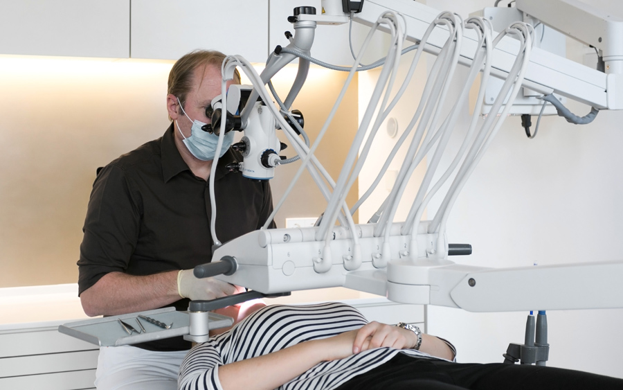 Dentysta z brązowej koszulki opracowuje zęba pacjentki pod mikroskopem. Pacjentka leży na unicie stomatologicznym xo care.