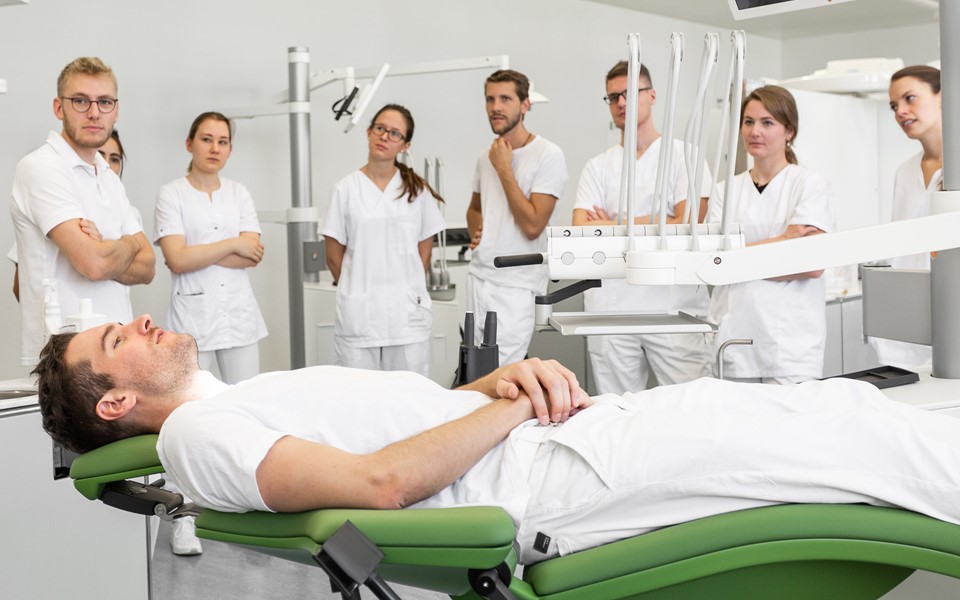 Studenci przyglądający się pacjentowi leżącemu na zielonym fotelu stomatologicznym